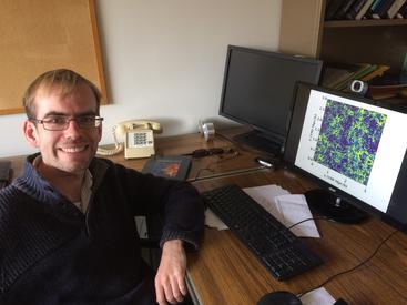 Dr. Simeon Bird poses, smiling in front of his computer